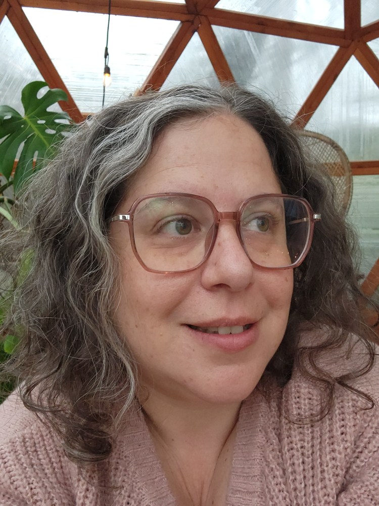 headshot photograph of Rebecca, a white woman with mid-length dark hair. She's wearing a pink sweater and glasses, and there's a green monstera plant behind her,